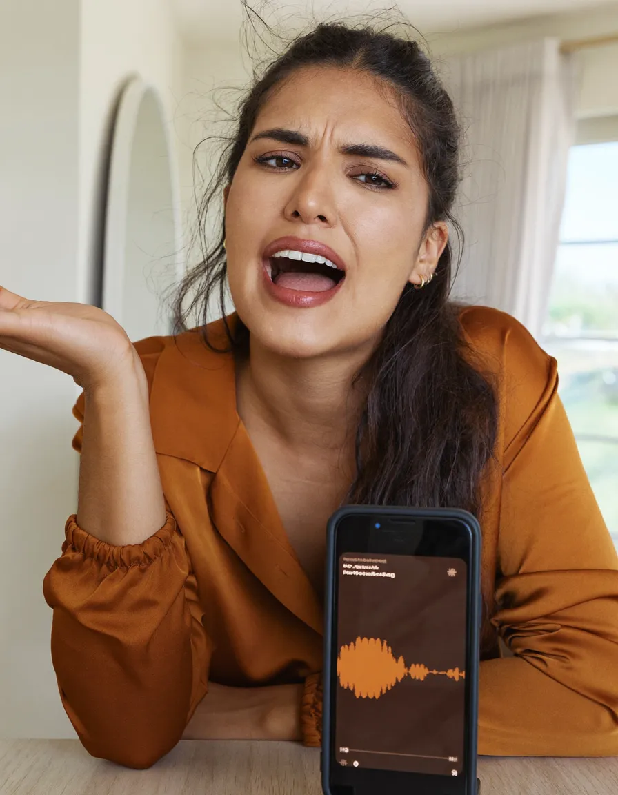 A woman sits at a table mid-sentence, gesturing as she speaks to a phone propped upright in front of her showing a live audio waveform.