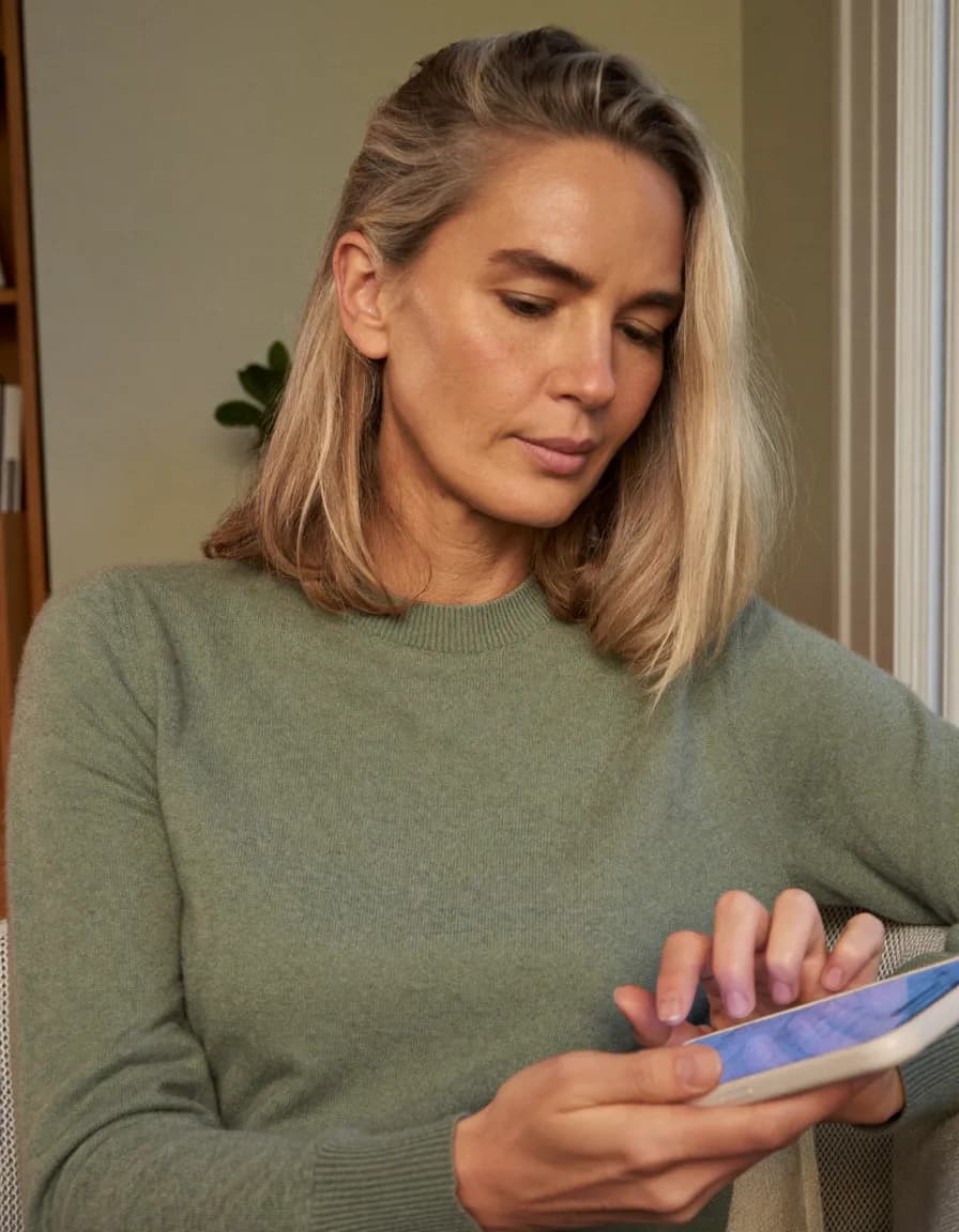 A woman sits alone in an armchair, looking down at the phone in her hands with a fingertip poised over the screen.