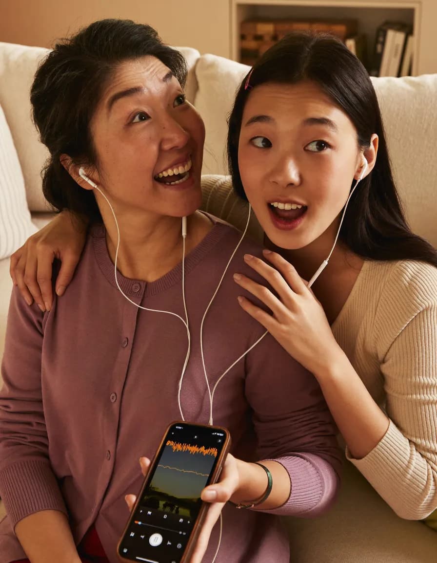 Two women sit close together on a sofa, sharing a pair of wired earbuds and smiling as a recording plays on the phone one of them holds.